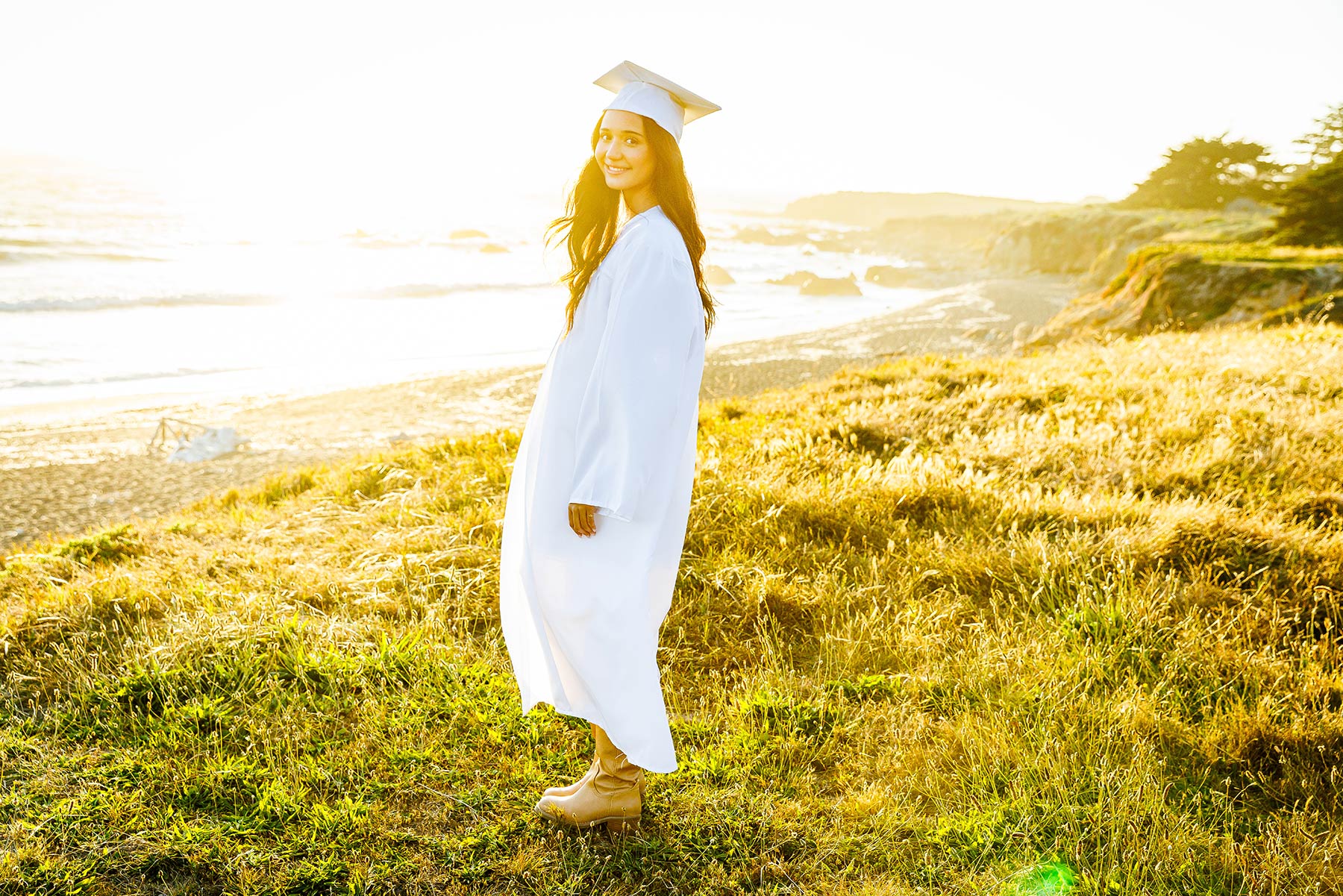 Wide-angle environmental senior portrait featuring a graduate in her gown standing on the bluffs overlooking the Pacific Ocean in Cambria.