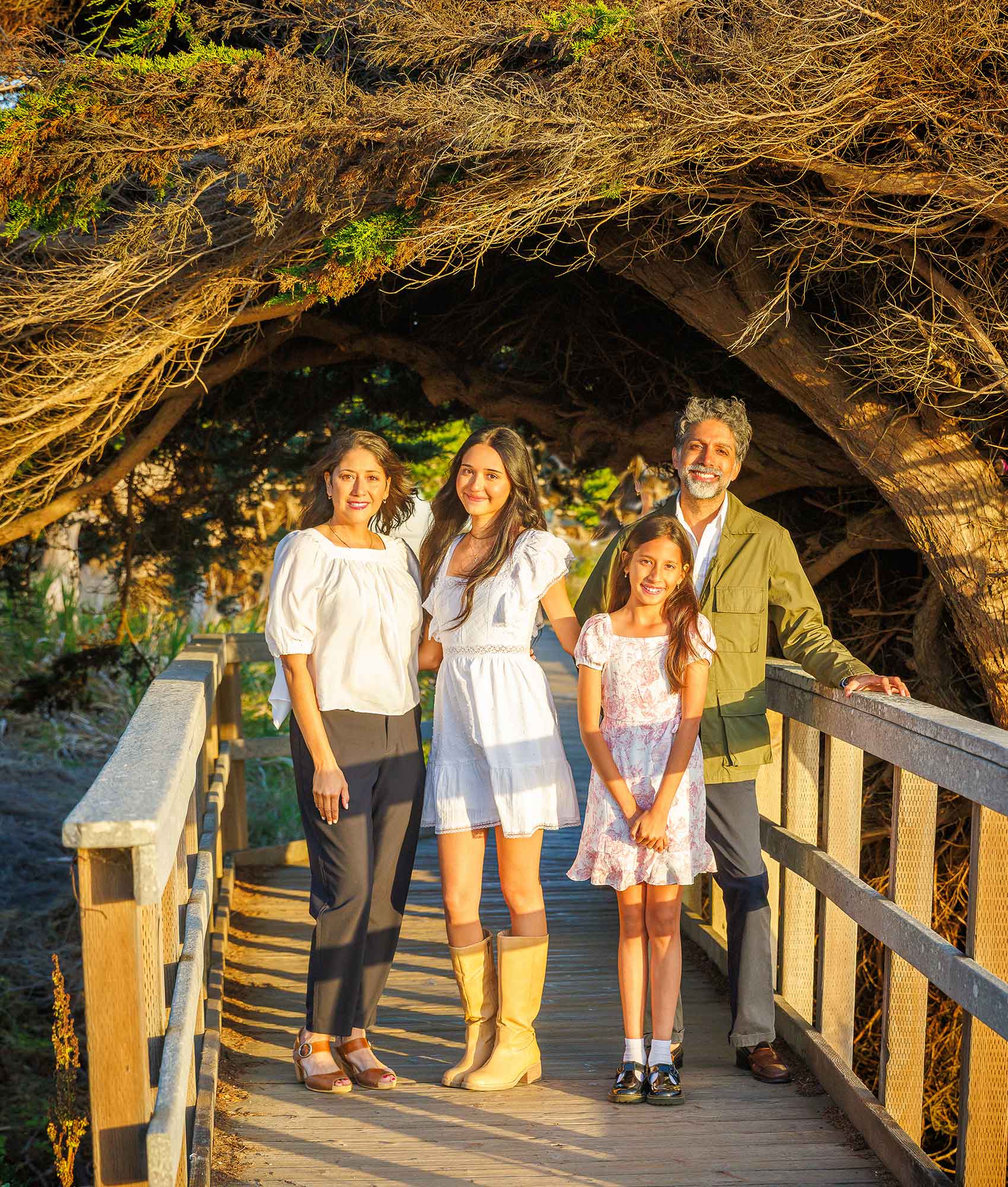 Family of four standing on a rustic wooden boardwalk under a cypress tree canopy during a Morro Bay portrait session.