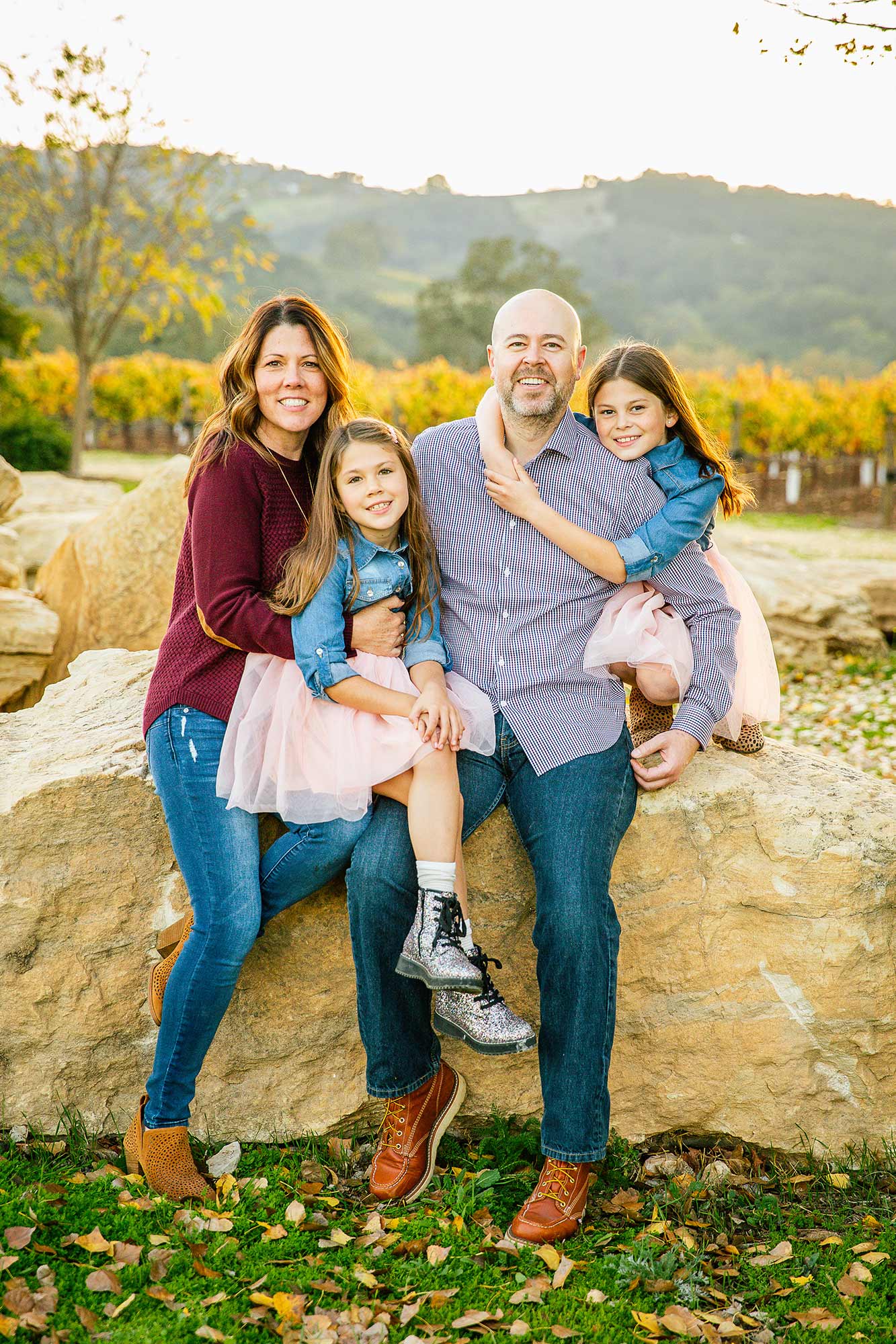 Natural light family portrait at a Paso Robles vineyard during autumn, featuring parents and two daughters sitting on large rocks with rolling hills in the background