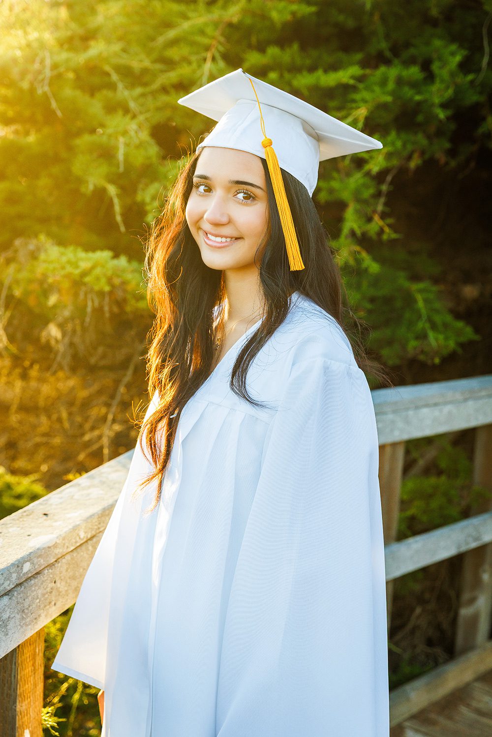 Classic graduation portrait of a senior in a white cap and gown on a wooden bridge at Moonstone Beach, Cambria.